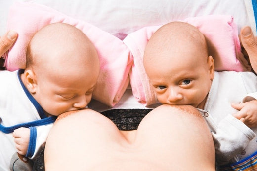 Mother breastfeeding twin babies who are lying on pink pillows, each nursing from one breast.