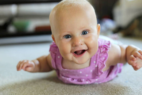 Smiling baby in a purple outfit lying on their stomach on a soft carpet, looking up excitedly.