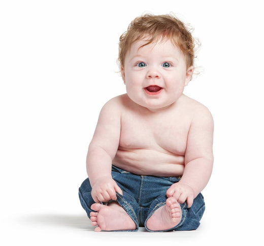 Smiling baby with curly hair sitting upright, wearing jeans, against a plain white background.