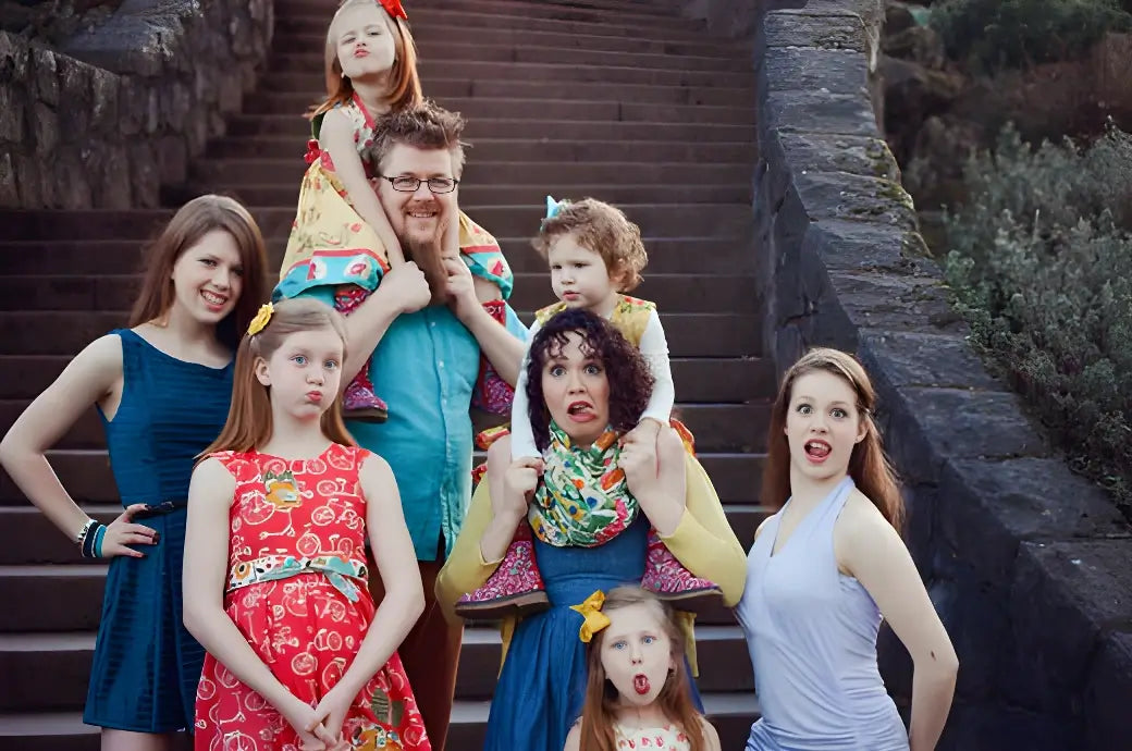 Family posing playfully on outdoor stone steps; two children sit on their parents' shoulders while others make funny faces around them.