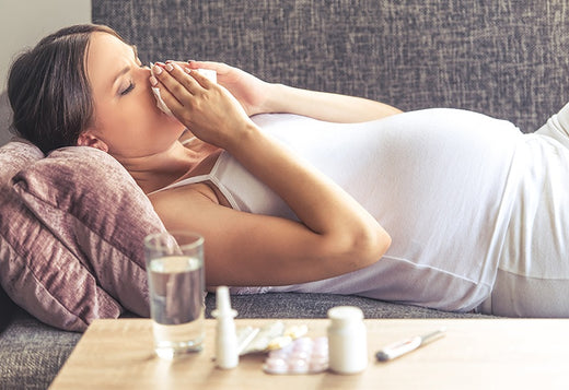 ant woman lying on a couch, holding a tissue to her nose, with a table nearby containing water, nasal spray, pills, and a thermometer.