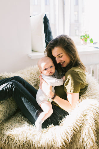 Smiling mother sitting in a cozy chair, holding her baby upright in a sunlit room.