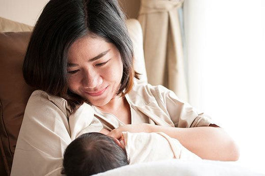 Smiling Asian mother breastfeeding her baby while sitting comfortably in a softly lit room.