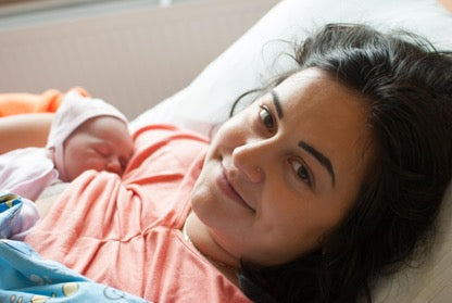 Smiling mother lying in a hospital bed with her newborn baby resting on her chest, both wrapped in blankets.