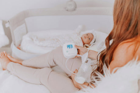 Mother sitting on a bed using a breast pump while looking at her baby lying in a bassinet nearby.