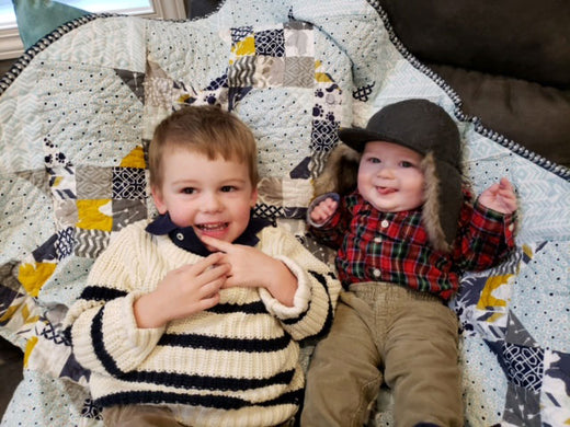 Two young siblings sitting on a quilted blanket; one wearing a cream and navy striped sweater, and the other dressed in a red plaid shirt with a cozy hat.