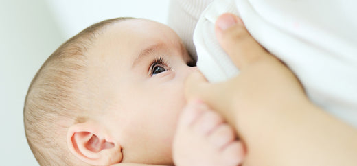 Close-up of a baby breastfeeding, gazing up while nursing against a soft, neutral background.