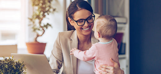 Smiling mother in glasses and a blazer holding her baby while working on a laptop at home.