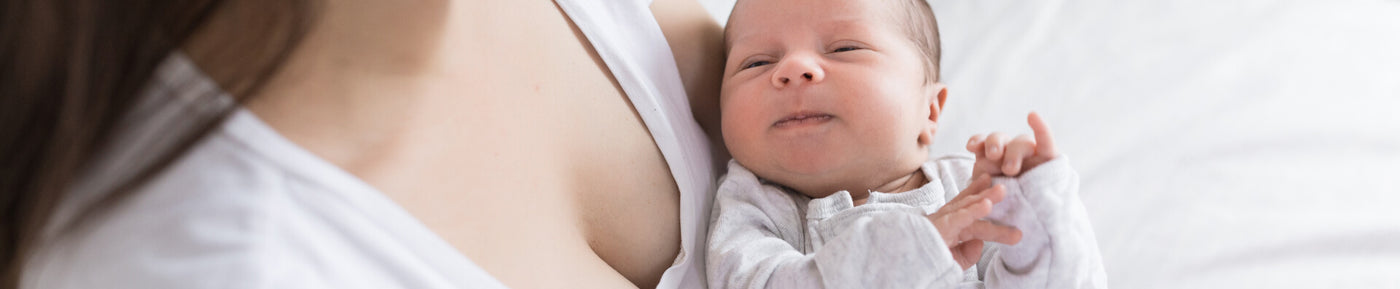 A close-up image of a woman holding a newborn baby against her chest. The baby, wearing a light gray outfit, has a slight smile and is looking up. The woman's neckline is visible, and her hair is partially out of view. The background is softly blurred, emphasizing the gentle and nurturing connection between the mother and baby.