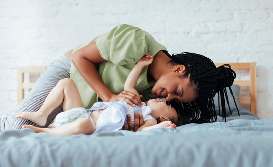 A mother and her baby are playing together on a bed. The mother, with braided hair and wearing a light green blouse, leans down towards her child, who is lying on their back. The baby, wearing a light blue shirt, reaches up with one hand while giggling. Their faces are close together, creating a joyful and intimate moment. The scene is cozy, with a neutral-colored bedding and a soft, inviting atmosphere. The background features a light brick wall, enhancing the warmth of the setting.