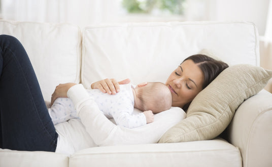 A mother gently cradles her sleeping baby in her arms while sitting on a light-colored sofa. The mother, dressed in a white top, has a soft smile as she gazes lovingly at her child, whose head rests against her shoulder. The baby is dressed in a light blue outfit with small patterns and appears relaxed. The setting is bright and airy, with soft natural light coming through the window, and the sofa contains a plush, textured cushion beside them.