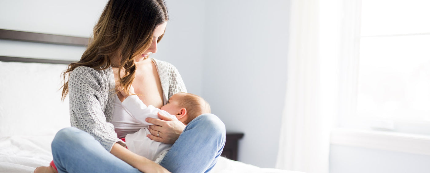 A woman with long, brown hair is sitting on a bed, breastfeeding an infant. She is wearing a light gray cardigan over a simple white top and light blue jeans. The baby, dressed in a white onesie, is cradled in her arm, facing her, while she gently holds the baby’s head with one hand. The scene is softly lit by natural light coming from a nearby window, creating a calm and intimate atmosphere. The background features pale blue walls and a white bedspread.