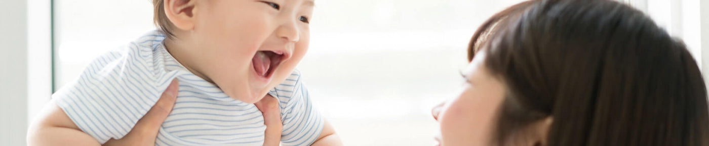 A close-up image of a joyful baby being held up by a woman. The baby, with short, light brown hair, has a big smile and is laughing, showing excitement. The woman, whose face is partially visible, looks at the baby with a warm smile, creating a loving interaction. The background features soft, blurred light coming through a window, emphasizing a bright and cheerful atmosphere. The baby is wearing a light blue and white striped shirt.