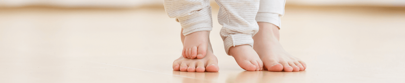 A close-up of two pairs of feet on a smooth wooden floor. On the left, a small child's foot is positioned next to an adult's foot on the right. The child's foot is slightly turned outward, while the adult’s foot is flat on the ground. Both feet are bare, showcasing soft, smooth skin. The background is softly blurred, with warm, natural lighting illuminating the scene.