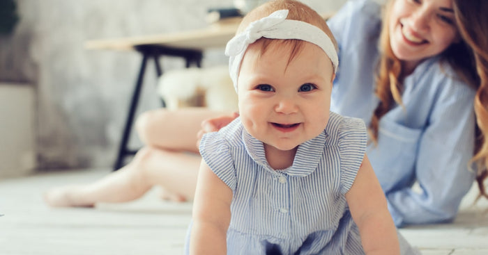 A smiling baby girl with blue eyes and a light blue and white striped dress crawls towards the camera. She wears a white headband with a bow, and her cheeks are slightly chubby, highlighting her happiness. In the background, a parent or caregiver with long hair is laughing and looking at the baby, seated on a light-colored floor. The setting has a cozy and warm atmosphere, with soft furnishings dimly visible.