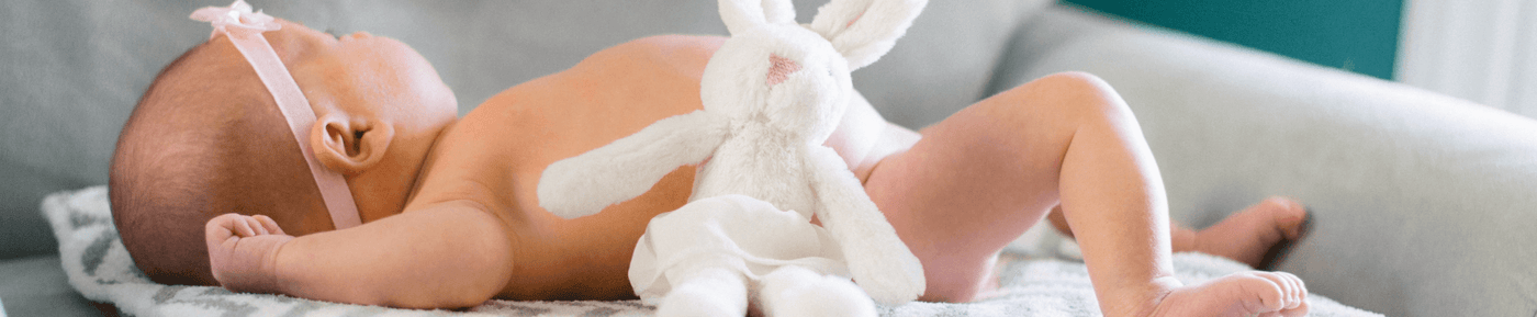 A close-up of a sleeping baby lying on their stomach on a soft, patterned blanket, with a white stuffed bunny resting against their back. The baby has a light pink headband adorned with a bow and is partially visible in a cozy indoor setting. The background is softly blurred, emphasizing the baby and the bunny.