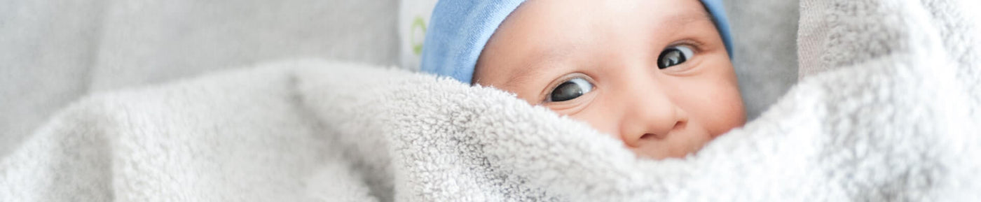 A close-up image of a baby peeking out from a soft, light gray blanket. The baby has a playful expression, with wide eyes and a slight smile, and is wearing a blue hat. The background is blurred but suggests a cozy indoor environment, creating a warm and inviting atmosphere.