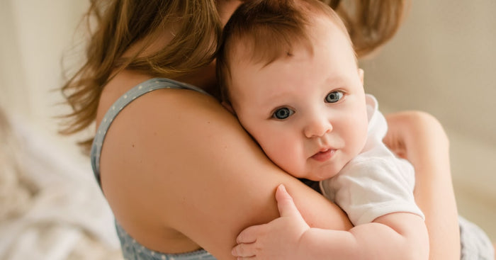 A close-up of a tender moment between a parent and a baby. The parent, with shoulder-length brown hair, is holding the baby tightly against their shoulder. The baby has light skin, bright blue eyes, and is wearing a simple white onesie. The baby's expression is curious and slightly pouting. The parent's shoulder is partially visible, showcasing a blue and white polka dot strap. The background is softly blurred with warm, neutral tones, creating a cozy atmosphere.