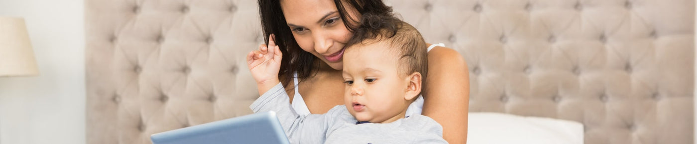 A mother sits on a bed, closely interacting with her baby who is nestled in her lap. Both are focused on a tablet in front of them. The mother, with dark hair, smiles gently as she points at the screen, while the baby, wearing a light gray shirt, appears curious and engaged, with one hand raised. The background features a softly designed fabric headboard and a bedside lamp, creating a warm and inviting atmosphere.