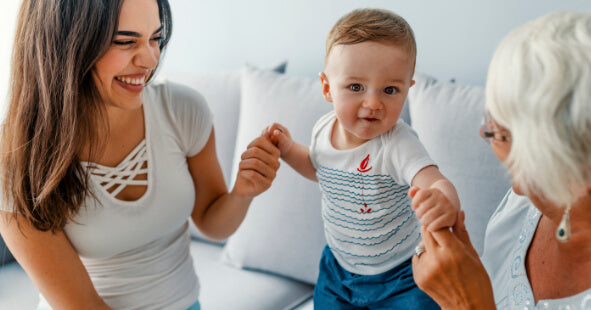 A joyful scene featuring a young woman and a grandmother interacting with a toddler. The woman, with long dark hair, is laughing and holding the child's hands, encouraging him to stand. The toddler, wearing a white shirt with blue stripes and denim shorts, looks delighted. The grandmother, with short gray hair and glasses, is smiling and reaching out to the child, creating a warm and loving atmosphere.