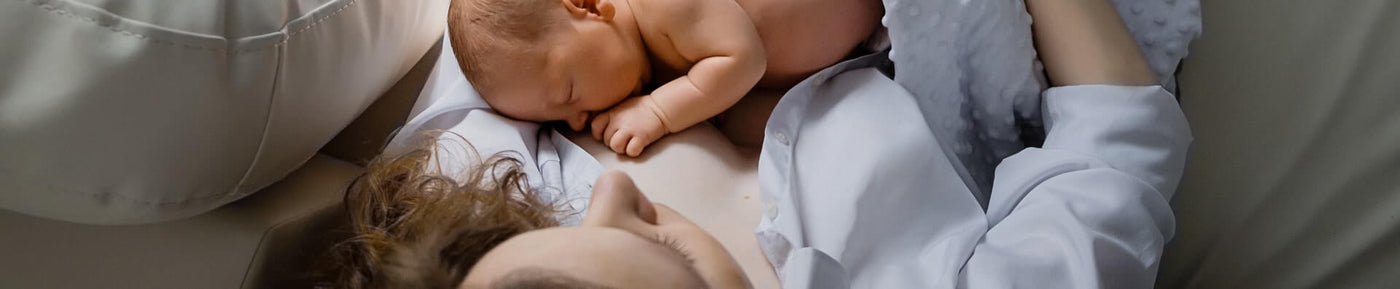 A close-up image of a woman lying down, gently cradling a newborn baby on her chest. The woman has curly hair, and she is wearing a light-colored shirt. The baby is curled up, sleeping peacefully with their head resting against the mother's skin. The background is soft and neutral, suggesting a cozy and intimate setting. The focus is primarily on the tender moment between the mother and child.