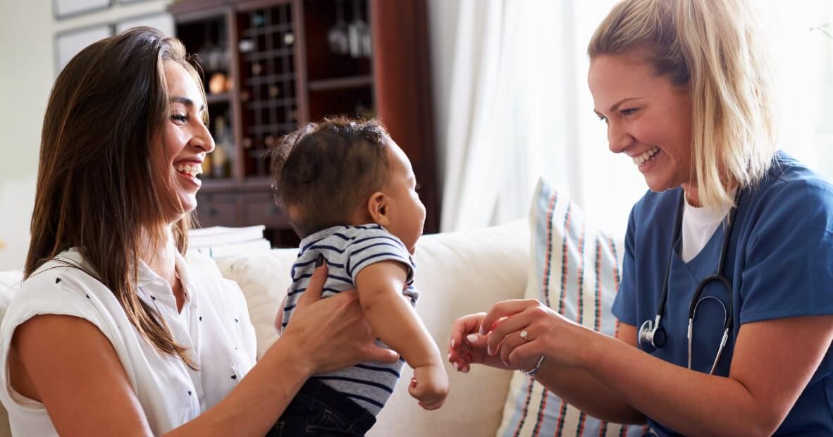 A smiling mother holds a baby facing away from the camera, while a cheerful healthcare professional sitting across from them interacts with the baby. The mother is dressed in a white blouse and has long, dark hair. The healthcare worker wears a blue medical uniform and has her hair tied back. The background features a softly lit room with a beige couch and a bookcase, creating a warm and inviting atmosphere. The women appear engaged in a friendly conversation, and the baby looks curious and happy.