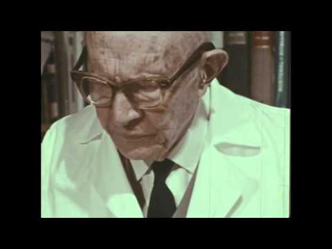 An elderly man wearing thick, rounded glasses and a white lab coat looks down thoughtfully. His face is wrinkled, indicating age and experience. He is situated in a library or study filled with books in the background. The image captures a moment of contemplation, emphasizing the seriousness of his demeanor.