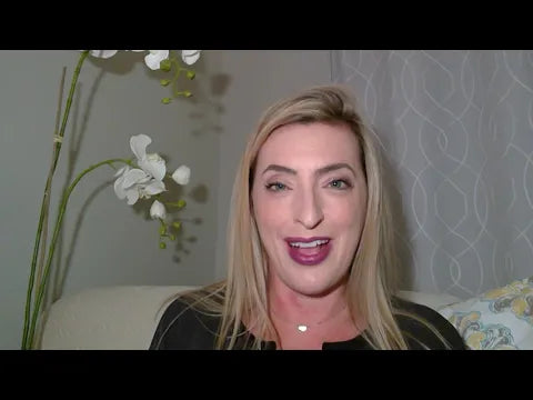 A woman with long, straight blonde hair smiles while speaking directly to the camera. She is wearing a black top and is seated on a light-colored couch. In the background, there is a decorative plant with white flowers and a patterned pillow. The room has soft lighting and a sheer curtain, creating a warm and inviting atmosphere.