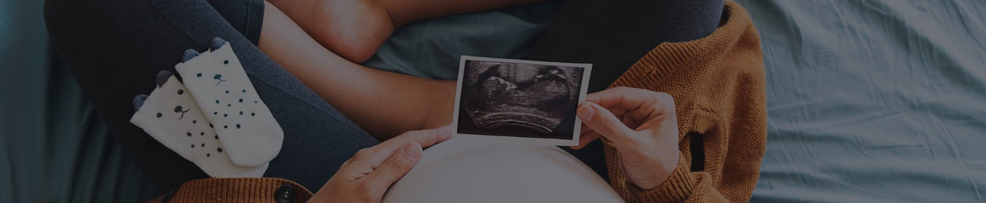 A person is sitting cross-legged on a bed, holding a black and white ultrasound photo in their hands, which displays a developing baby. They have a round belly visible, and are wearing a cozy brown sweater. On the floor nearby, a pair of white infant socks with cute bear designs is seen. The background consists of soft bedding and blankets in muted colors, creating a warm and nurturing atmosphere.