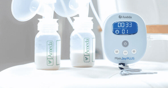 A close-up image of a breast pump setup. Two clear baby bottles, each filled with expressed milk, are positioned on a flat surface. The bottles feature a green logo with the brand name "Ameda." Beside the bottles is a breast pump control unit labeled "Mya Joy PLUS," which has a digital display showing a timer reading 00:33 and a numerical display with the number 01. The unit has various buttons for operation, and the background is softly lit, creating a clean, clinical appearance.