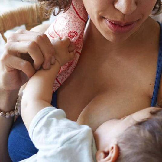 A mother is breastfeeding her baby while gently holding the baby's hand. The mother's face is partially visible, showing a calm expression, and she is wearing a blue top. The baby, whose head is turned towards the mother's chest, is wearing a light-colored shirt. A patterned fabric, possibly a nursing scarf, is draped around the mother's neck. The background features soft lighting and hints of a cozy setting.