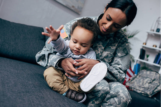 A female soldier in a camouflage uniform sits on a couch, lovingly holding her young child. The child, wearing a gray hoodie and brown pants, reaches out with one hand while kicking a foot forward. An American flag patch is visible on the soldier's sleeve, and military gear is seen in the background.