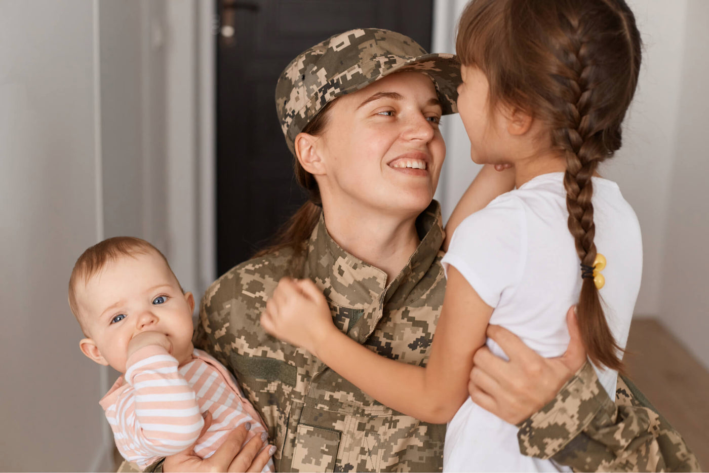 A female soldier in a camouflage uniform smiles while embracing her two young children. She holds a baby in a striped outfit sucking on their hand and a little girl with braided hair wearing a white shirt. They are indoors, sharing a warm and loving moment.