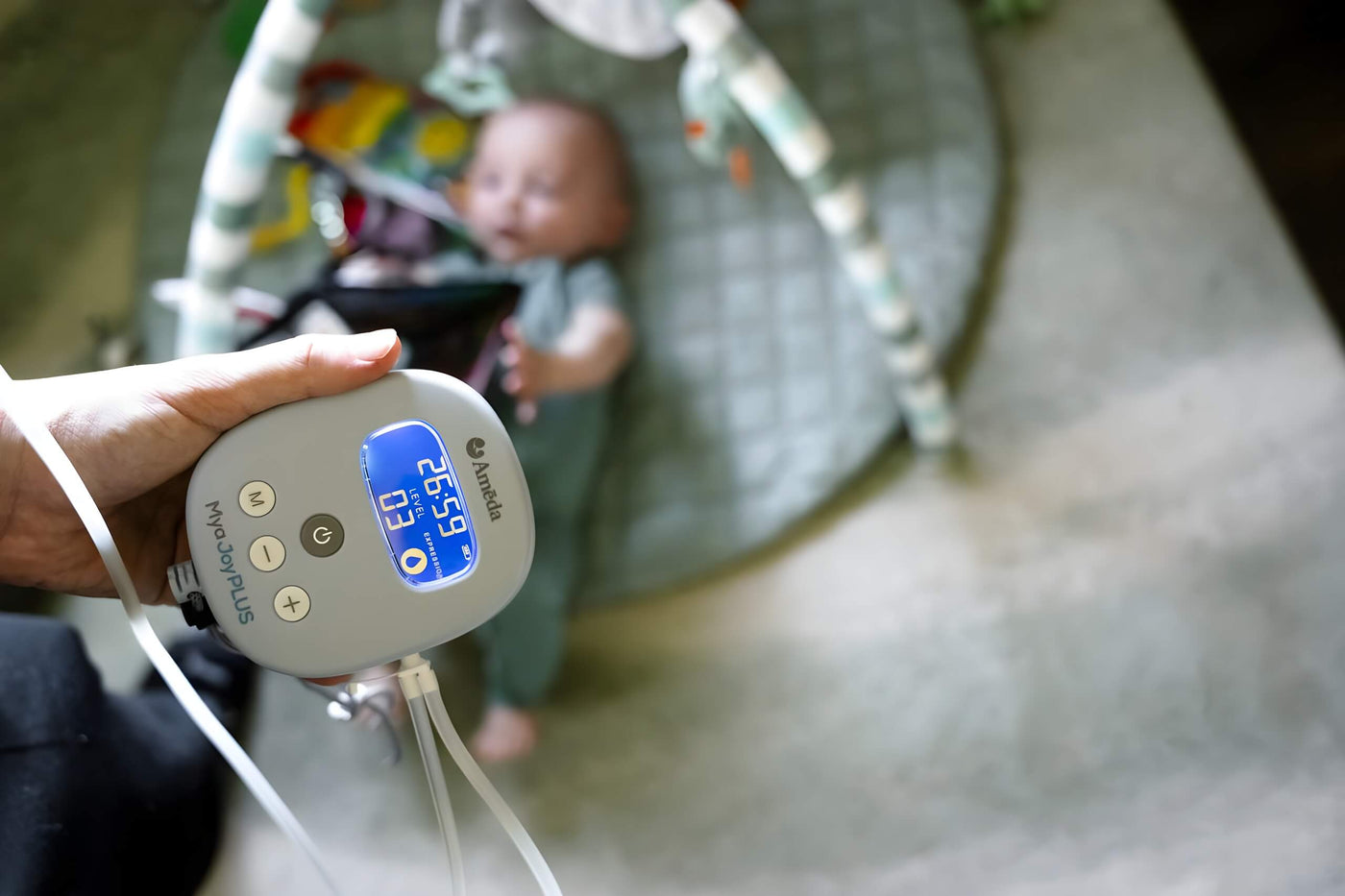 A person holding an Ameda Mya Joy Plus Breast Pump device with a digital screen displaying settings, while a baby lies on a play mat in the background.