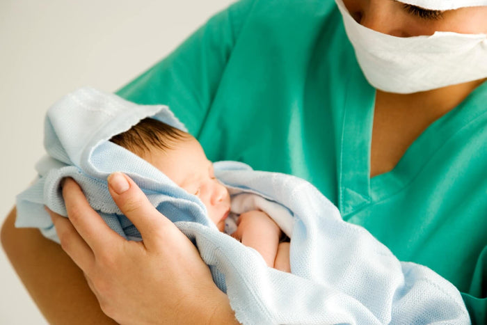 Healthcare professional in scrubs and a mask gently holding a newborn baby wrapped in a light blue blanket.