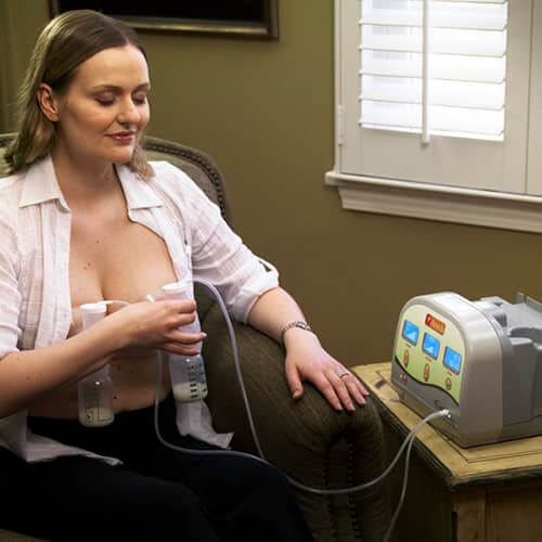 Mother seated in a comfortable chair using a hospital-grade Ameda breast pump, holding the flanges against her chest while expressing milk.