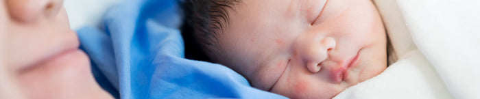 A close-up image of a sleeping newborn baby, nestled against a soft white blanket. The baby has dark hair and closed eyes, with a peaceful expression. The baby's mouth is slightly open, resting against a person's cheek, which is partially visible on the left side of the image, revealing smooth skin and a subtle pink hue. The background is softly blurred, providing a serene atmosphere to the moment.