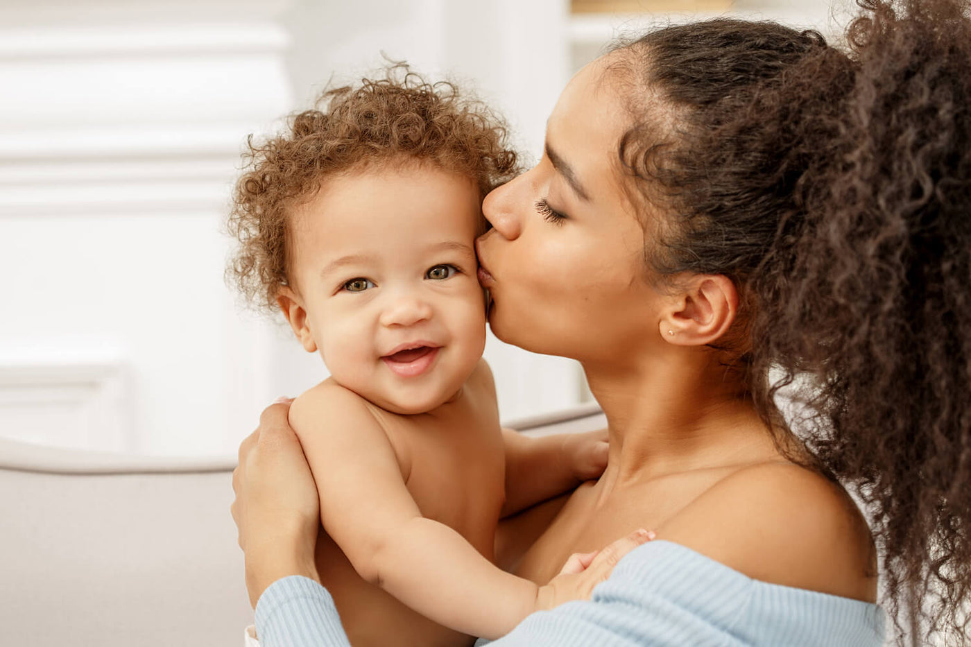 A woman with curly hair gently kisses a smiling baby with curly hair on the cheek. The baby is being held in her arms, facing the camera with a joyful expression. The setting is bright and airy, featuring soft, neutral tones in the background. The woman is wearing a light blue sweater, while the baby is bare-chested. The mood of the image is warm and affectionate.