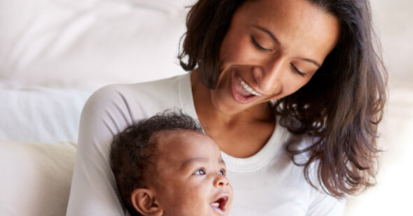 A woman with medium skin tone and long, wavy black hair is smiling joyfully at a laughing baby being held in her arms. The baby, with curly hair and a light brown complexion, looks delighted and is gazing back at the woman. They are seated on a soft, light-colored couch in a bright, cozy setting. The woman's expression radiates warmth and affection as they share a playful moment together.