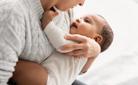 A close-up image of a caregiver holding a baby. The caregiver, wearing a light-colored knitted sweater, is looking down at the baby with a gentle expression. The baby, dressed in a white onesie, is gazing back up at the caregiver, displaying a curious and joyful demeanor. The background is softly blurred and appears to be a light-colored bed or surface, creating a calm and intimate atmosphere. The image conveys warmth and tenderness in their interaction.