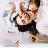 A joyful mother lies on a bed, laughing while holding her baby who is nestled against her shoulder. The baby, wearing a light gray onesie, is reaching out to touch the mother's face. The mother has long, dark hair and is dressed in a white top, radiating happiness. In the foreground, there is a box labeled "Ameda Spare Parts Kit," featuring images of components for breast pumps, with text in English and French. The background shows a light-colored, textured blanket.