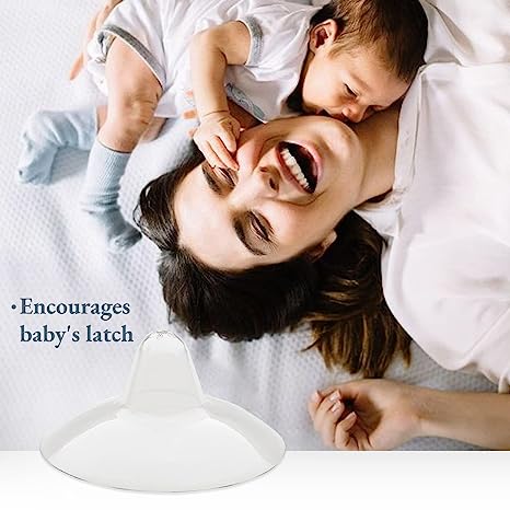 A joyful mother smiles widely while holding her baby, who is playfully grasping her face. The mother has long, dark hair and is lying on a light-colored bed with textured bedding. In the foreground, there is a white breastfeeding accessory designed to support proper latching. To the left of the image, there is text that reads, "Encourages baby's latch." The overall mood of the image is warm and affectionate, highlighting the bond between the mother and child.