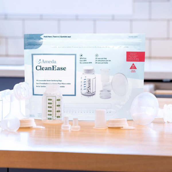 A package of Ameda CleanEase microwavable steam sanitizing bags is displayed in the center. The package is predominantly blue and white, featuring the Ameda logo and information about the product, including "BPA Free" and instructions for use. Surrounding the package are various white plastic accessories typically used for baby feeding or pumping, including bottle tops, adapters, and containers. The items are arranged on a light wooden surface with a blurred background of a kitchen setting.