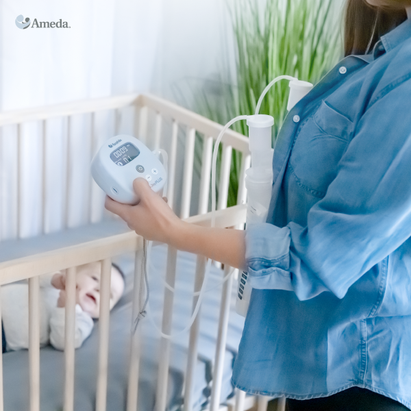 A woman in a light blue shirt stands next to a crib, holding a breast pump in one hand. The pump features a digital display showing the time. Inside the crib, a baby with light-colored hair is looking up with a curious expression. The background includes soft natural light and greenery, adding a serene atmosphere to the scene.