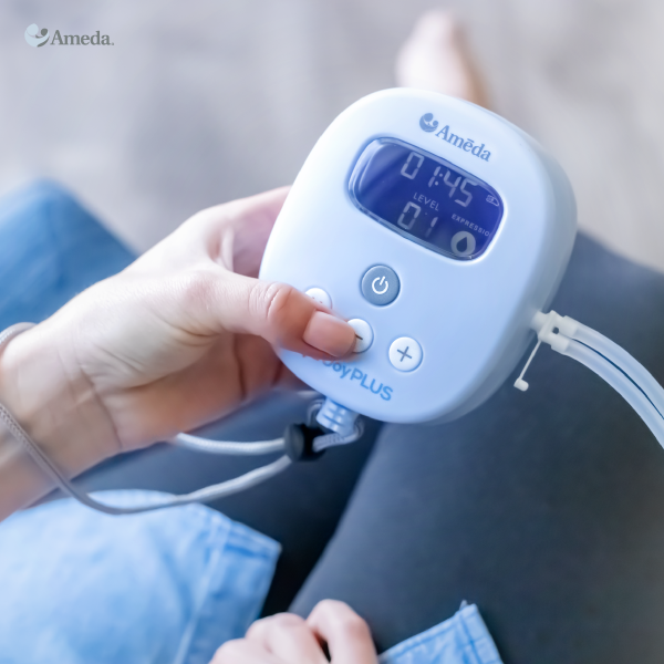 A close-up of a hand holding a blue breast pump controller with digital display. The display shows a time of 01:45 and a level of 01 for the expression setting. The background features a person's legs resting on a soft surface, with a denim fabric visible. The overall scene suggests a serene and personal setting for using the breast pump.