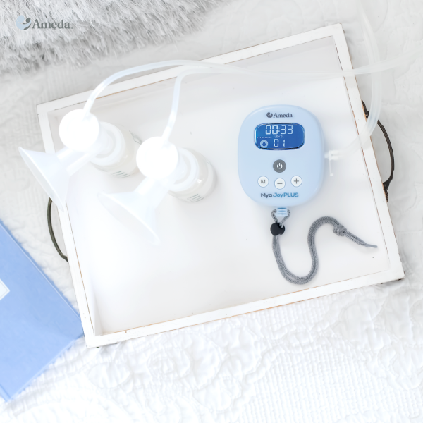 A flat, white tray on a soft, textured surface holds a blue digital breast pump control unit labeled "mya Joy PLUS" with buttons and a timer display showing "00:33." Two white breast shields are connected to the pump via clear tubes, placed next to two small white storage bottles. A partially visible blue box is in the background. The overall scene is bright and clean, emphasizing the breast pump setup.