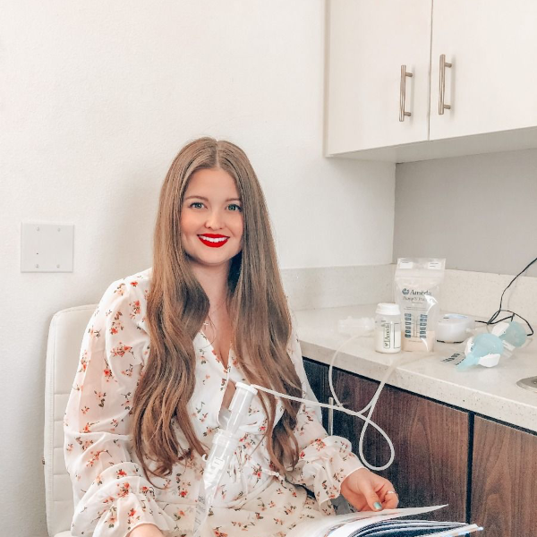 A young woman with long, wavy brown hair sits in a bright, modern room. She is wearing a white dress adorned with small floral patterns and has bright red lipstick. In front of her is a small table with various medical supplies, including a nebulizer connected to a tube that she is holding. The background features light-colored cabinetry, and she appears to be engaged in a relaxed activity, possibly reading or waiting for her treatment.