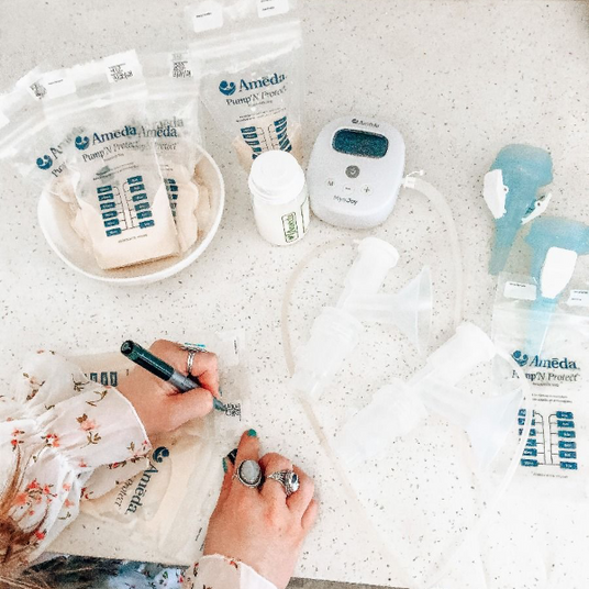 A flat surface displays various breastfeeding accessories and supplies. In the center, a person’s hand is holding a black pen, writing on a clear plastic bag labeled "Ameda Pump ‘N Protect." Surrounding this, there are several similar bags, white containers, and a small electronic device with a digital display. Two white breast pump flanges, a pair of blue flanges, and connecting tubes are also arranged on the surface.