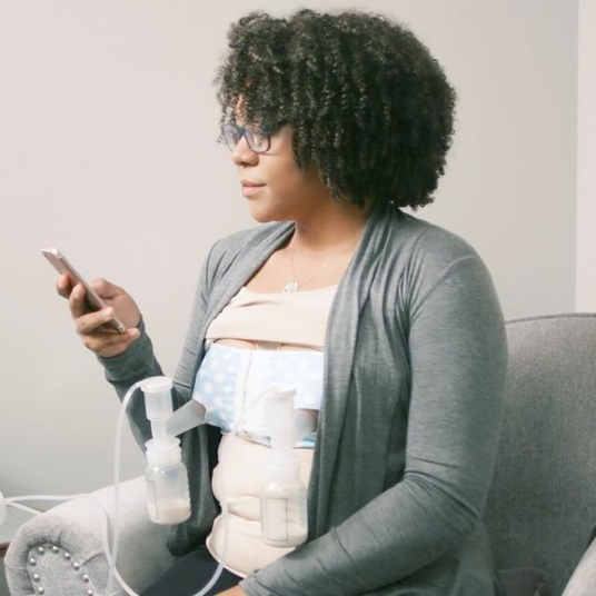 A woman with curly hair, wearing glasses, sits comfortably in a chair. She is holding a smartphone in her left hand while breast pumping. The pumping device is attached to her body with two bottles collecting milk, which are positioned on her chest beneath a light blue and white polka-dot shirt. She appears focused and relaxed in a softly lit room with neutral-colored walls.