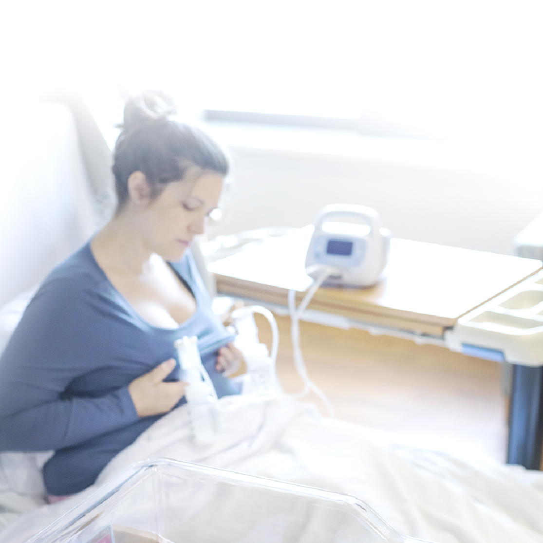 A woman sitting in a hospital bed is using a breast pump. She is dressed in a blue top and has her hair tied back in a bun. The atmosphere appears bright, suggesting sunlight coming through a nearby window. On a table beside her, there is a breast pump and two small bottles positioned for use. A transparent bassinet is visible in the foreground, and the woman is focused on her task, demonstrating a moment of quiet concentration. The bedding is white, adding to the serene hospital setting.